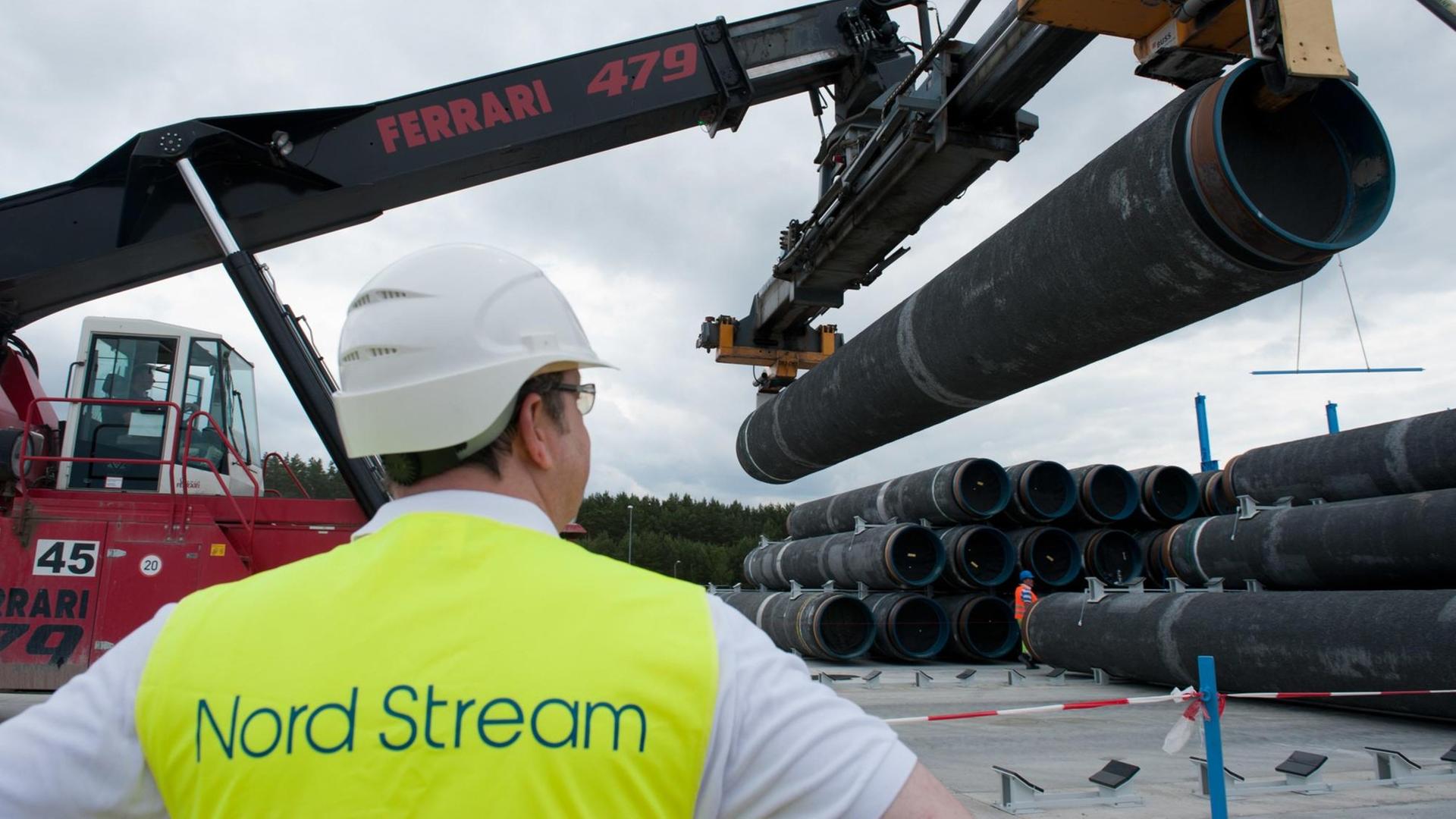 Im Vordergrund steht ein Mann in gelber Warnweste und beobachtet einen Bagger, der Pipelinerohre hebt. Im Vordergrund steht ein Mann in gelber Warnweste und beobachtet einen Bagger, der Pipelinerohre hebt.