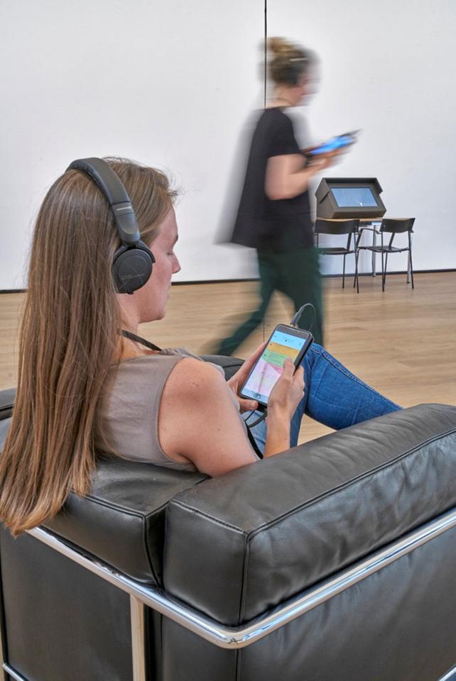 Installationsansicht "Radiophonic Spaces" im Museum Tinguely, Basel. Eine Frau sitzt in einem Sessel, hat einen Kopfhörer auf und schaut auf ihr Smartphone. Im Hintergrund geht eine Frau, ebenfalls einen Kopfhörer auf und schaut auf ihr Smartphon Installationsansicht "Radiophonic Spaces" im Museum Tinguely, Basel. Eine Frau sitzt in einem Sessel, hat einen Kopfhörer auf und schaut auf ihr Smartphone. Im Hintergrund geht eine Frau, ebenfalls einen Kopfhörer auf und schaut auf ihr Smartphon