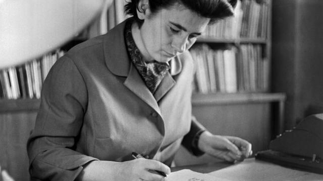 Christa Wolf sitzt 1963 vor einem Bücherregal an ihrem Schreibtisch und liest in einem Buch.