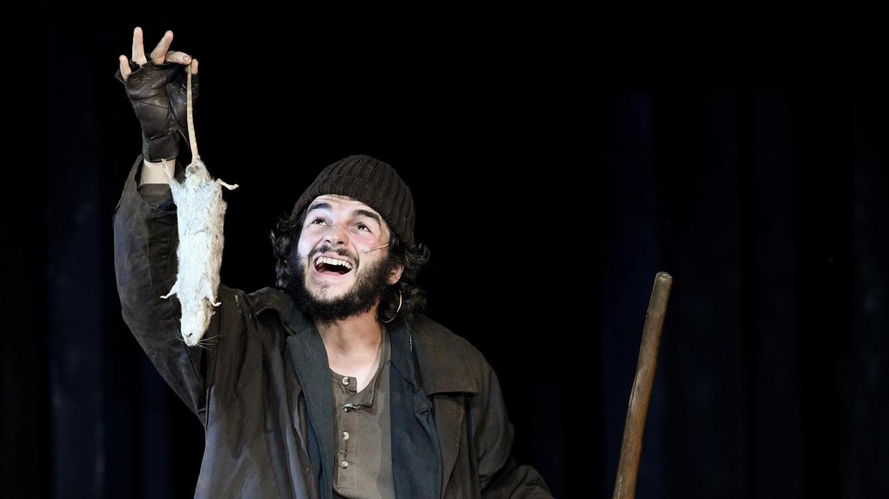 Der Schauspieler Rochus Rückel steht auf der Theaterbühne in einem Kostüm, das die Armut seiner Figur zeigt und hält eine tote Ratte am Schwanz haltend in die Höhe.