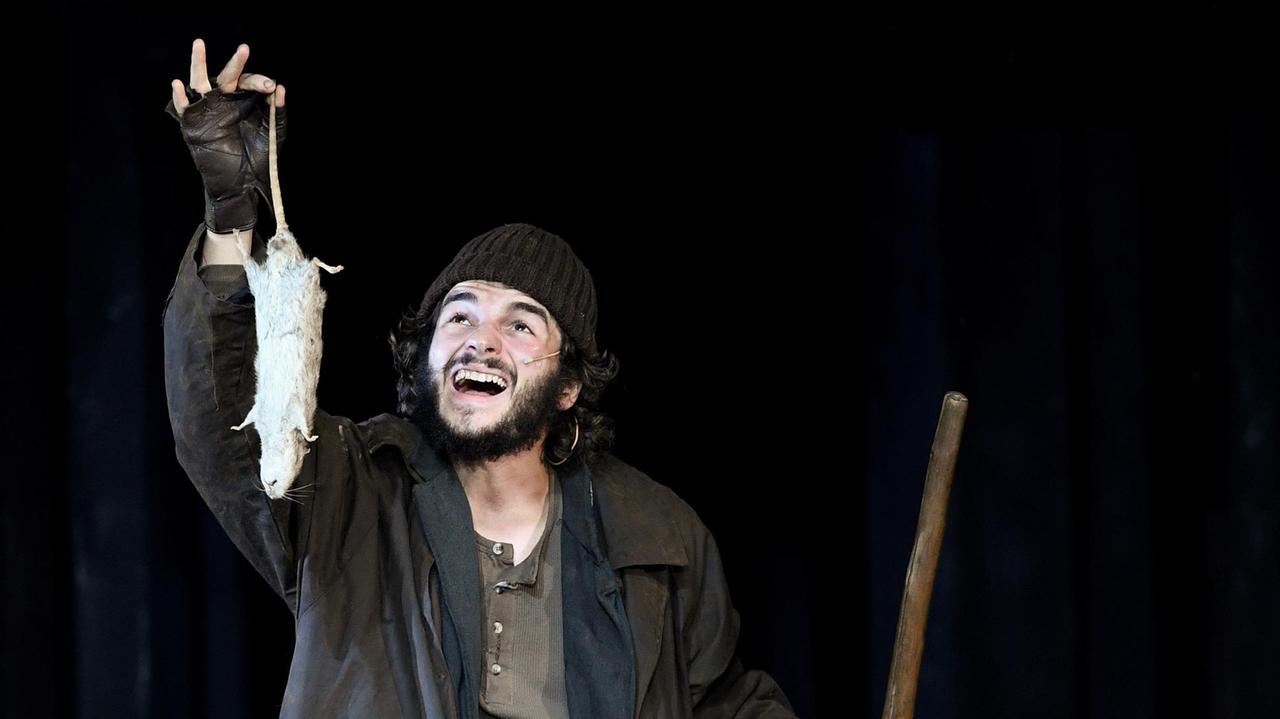 Der Schauspieler Rochus Rückel steht auf der Theaterbühne in einem Kostüm, das die Armut seiner Figur zeigt und hält eine tote Ratte am Schwanz haltend in die Höhe. Der Schauspieler Rochus Rückel steht auf der Theaterbühne in einem Kostüm, das die Armut seiner Figur zeigt und hält eine tote Ratte am Schwanz haltend in die Höhe.