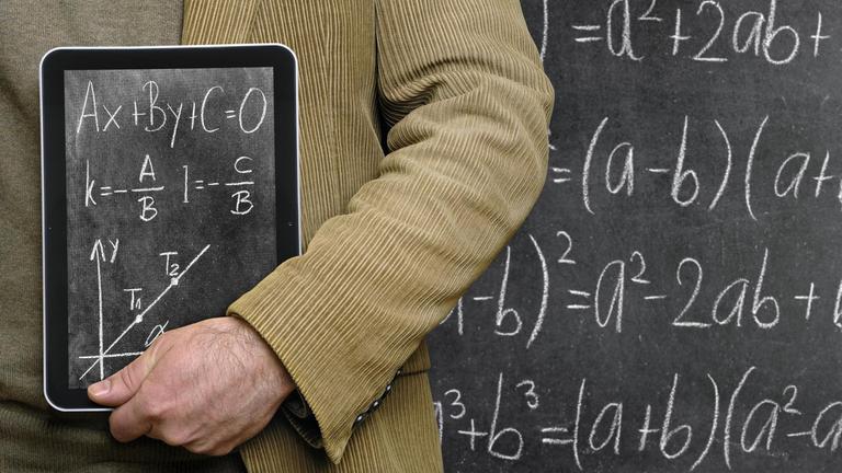 Lehrer hält einen Tablet-Computer vor einer Tafel, auf dem eine Tafel abgebildet ist. 