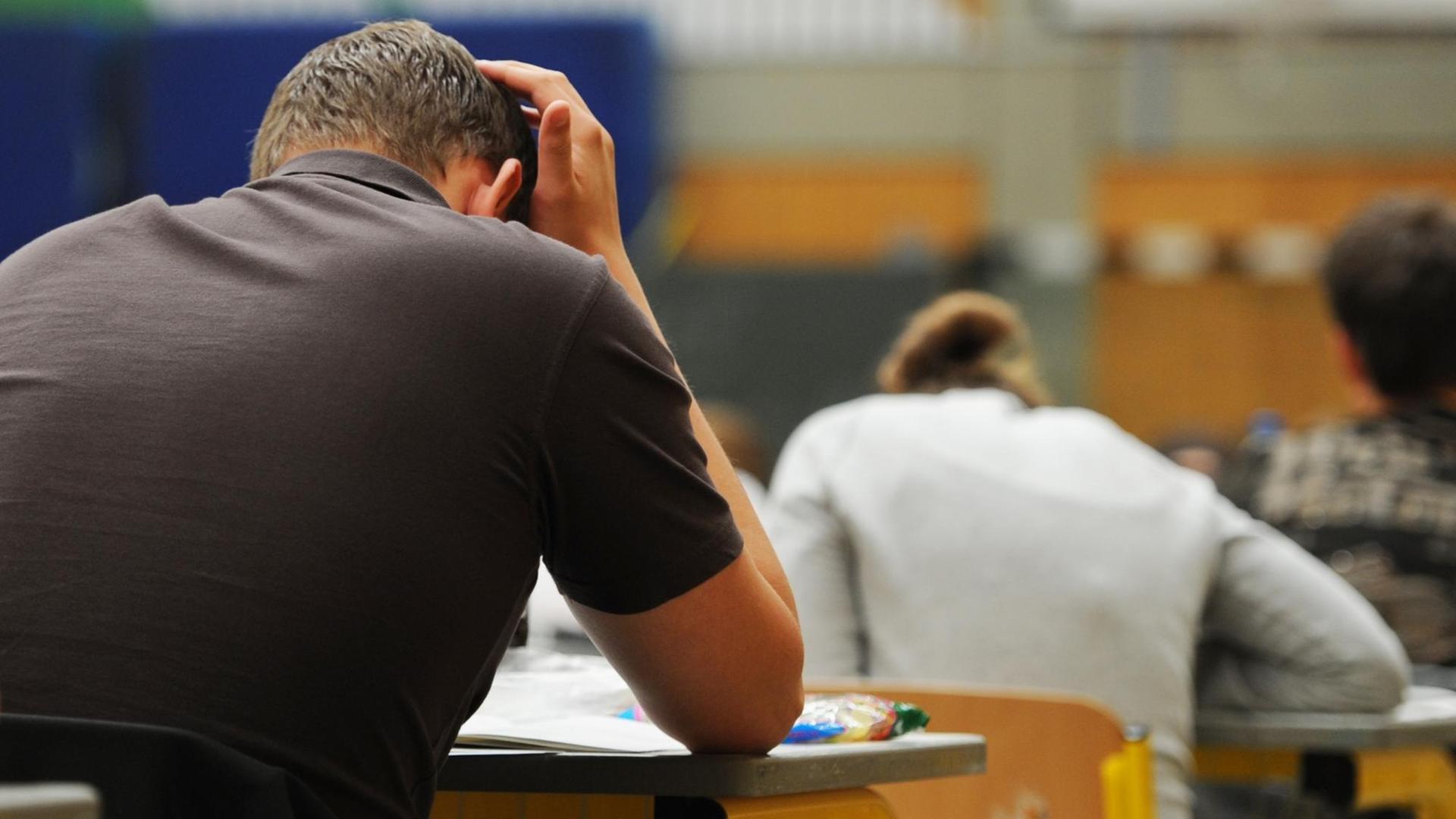 Zwei von schräg hinten aufgenommene Schüler sitzen während der Abiturprüfung an ihren Tischen in nachdenklicher Pose. Zwei von schräg hinten aufgenommene Schüler sitzen während der Abiturprüfung an ihren Tischen in nachdenklicher Pose.