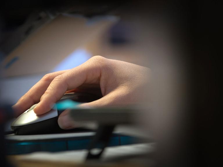 Eine Hand mit Computermaus wird durch eine Lücke in der Büroeinrichtung hindurch beobachtet.