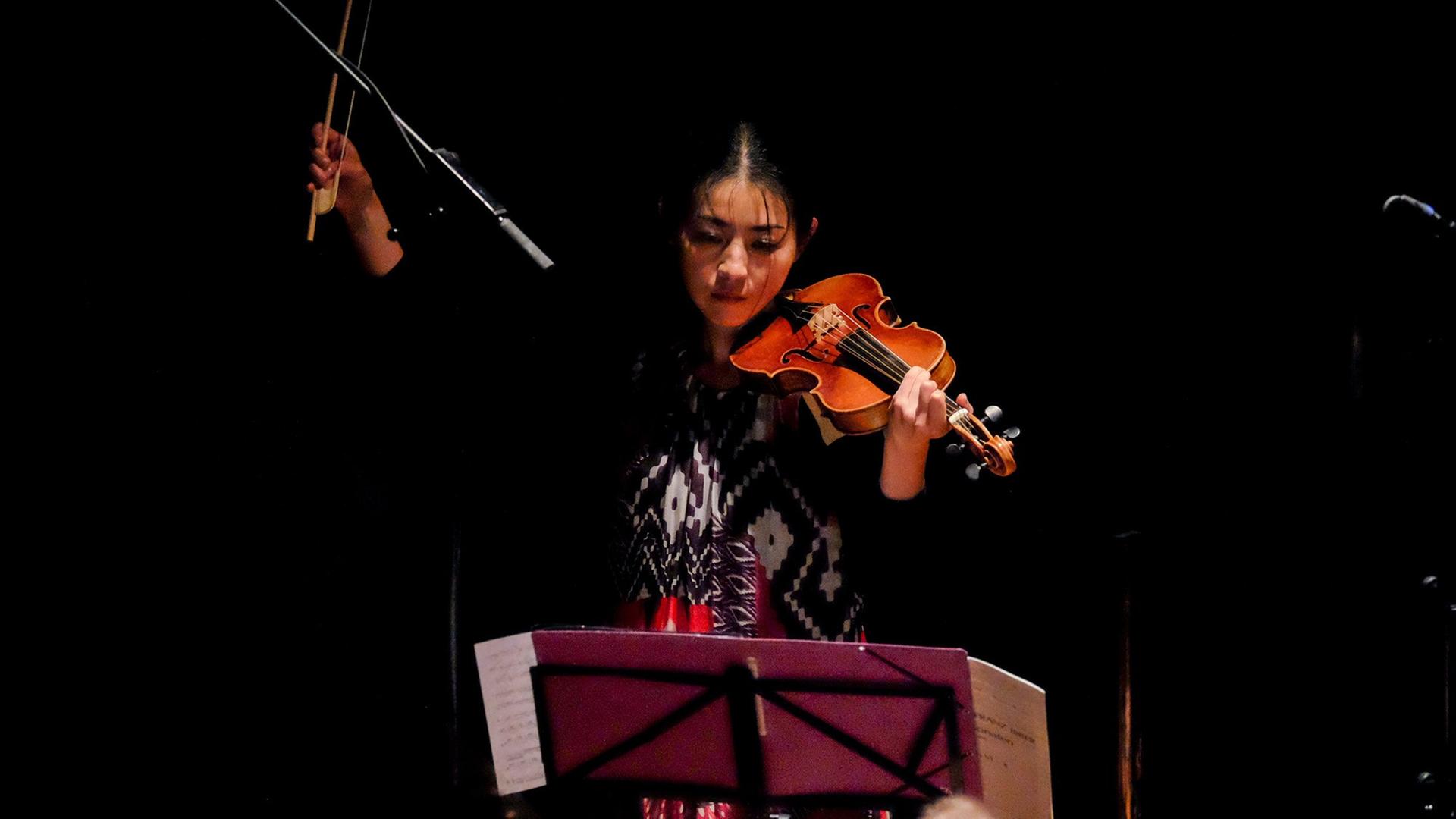 Die Geigerin Mayumi Hirasaki beim Spiel, den rechten Arm mit dem Bogen in der Hand hoch erhoben, umgeben von Schwarz. Die Geigerin Mayumi Hirasaki beim Spiel, den rechten Arm mit dem Bogen in der Hand hoch erhoben, umgeben von Schwarz.