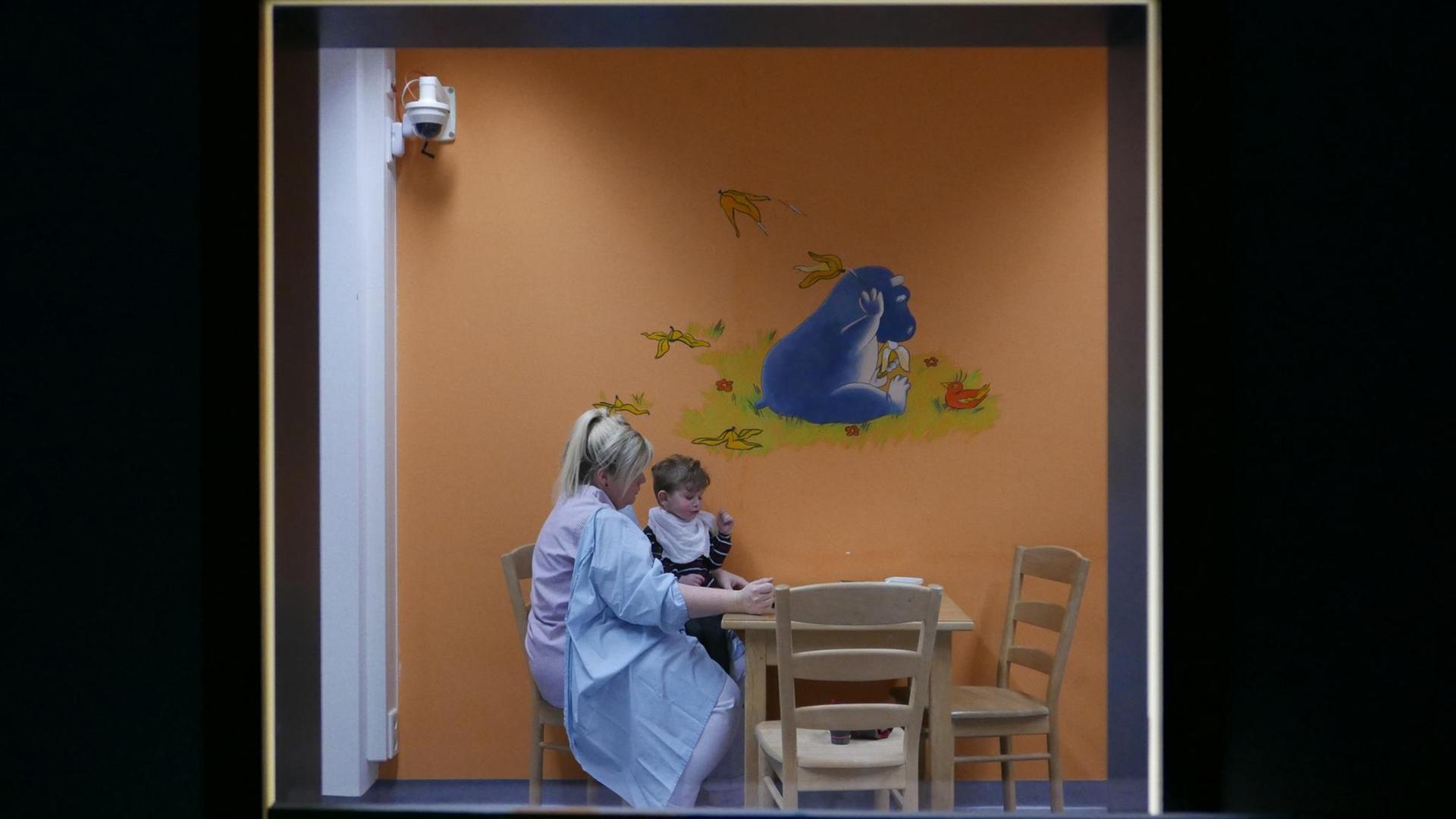 Durch eine Scheibe ist eine Klinikmitarbeiterin zu sehen, die mit einem kleinen Jungen am Tisch sitzt.