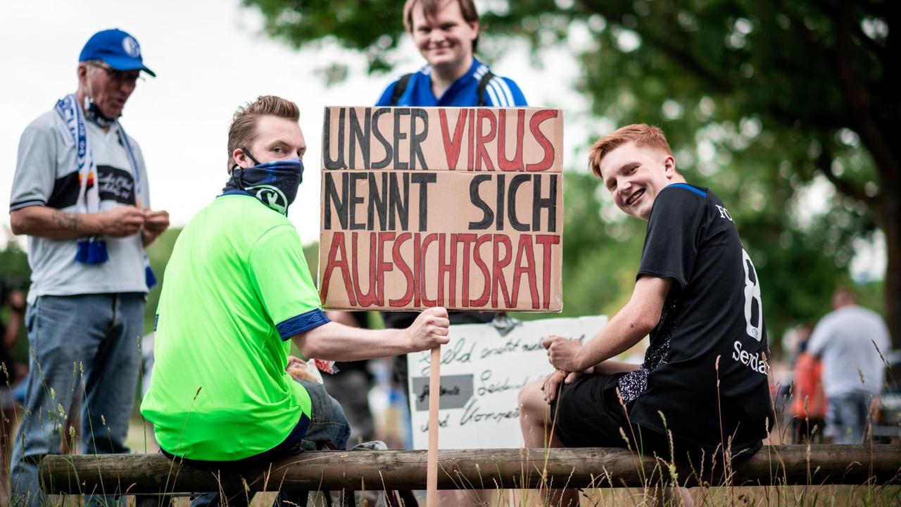 Zwei junge Männer sitzen auf einem Balken und halten ein Schild mit der Aufschrift "Unser Virus nennt sich Aufsichtsrats" in die Höhe. Dahinter weitere Demonstranten.