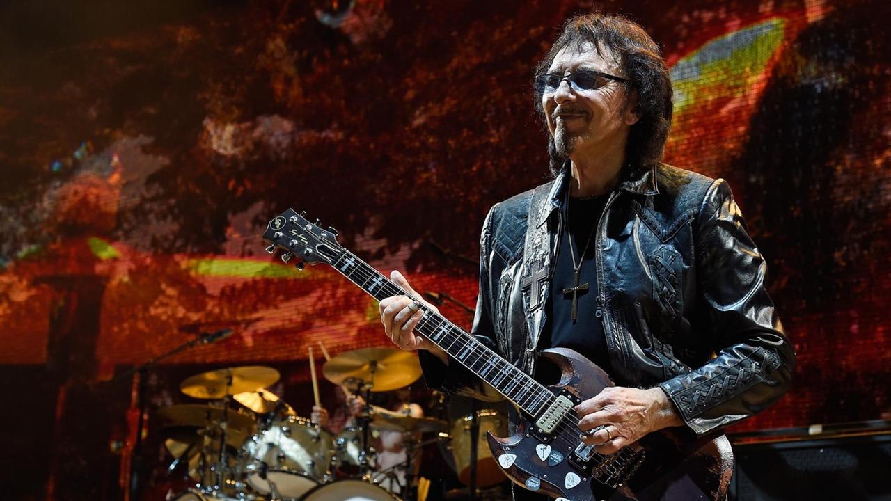 Gitarrist Tony Iommi von der Band Black Sabbath während eines Konzertes in Jones Beach. Er spielt auf seiner Gitarre, im Hintergrund steht das Schlagzeug und es werden düstere Bilder auf eine Leinwand projiziert.