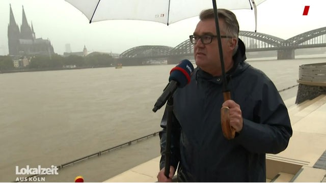 Christian David steht mit Schirm und Mikrofon am Rheinufer, im Hintergrund der Kölner Dom