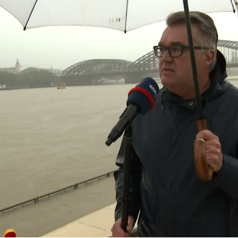 Christian David steht mit Schirm und Mikrofon am Rheinufer, im Hintergrund der Kölner Dom