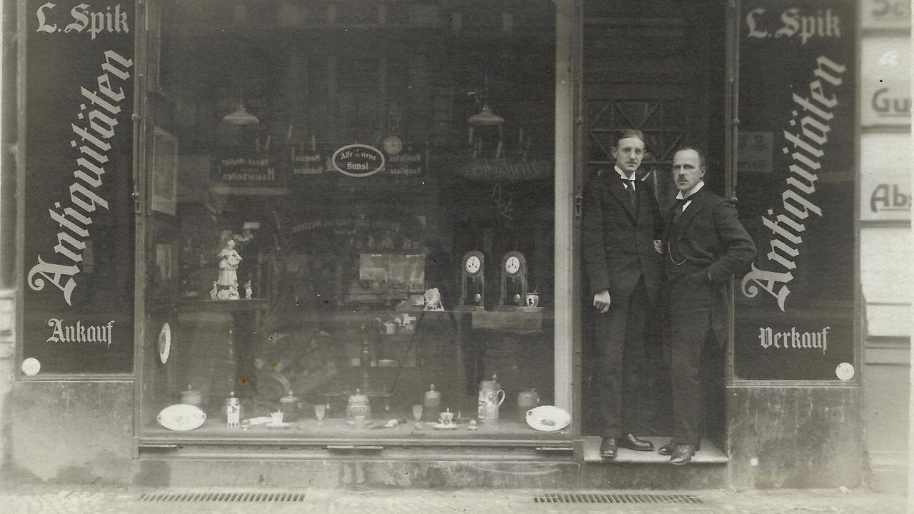 2 Männer auf einer historischen Aufnahme von 1919 vor einem Geschäft.