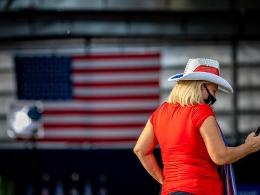 Eine Frau mit Cowboyhut schaut auf ihr Smartphone, im Hintergrund ist die amerikanische Flagge zu sehen.