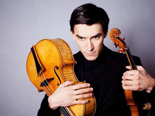 Der Geiger Sergey Malov hält in der einen Hand eine Violine und in der anderen ein Violoncello da spalla