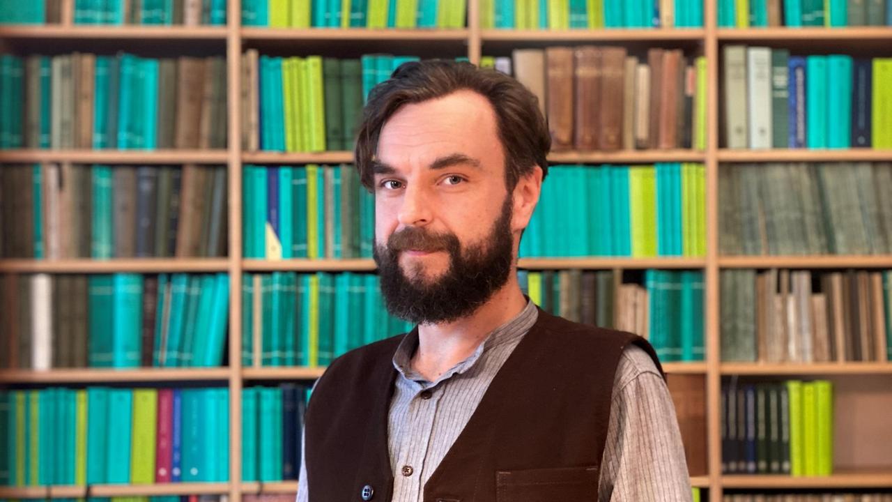 Ein lächelnder Mann mit Vollbart und in Weste steht vor einem Bücherregal. Ein lächelnder Mann mit Vollbart und in Weste steht vor einem Bücherregal.