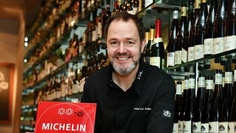 Der Sternekoch Marco Müller in seinem Restaurant Rutz. Er hält einen Michelin-Restaurantführer in Händen und blickt lächelnd in Richtung des Betrachters.