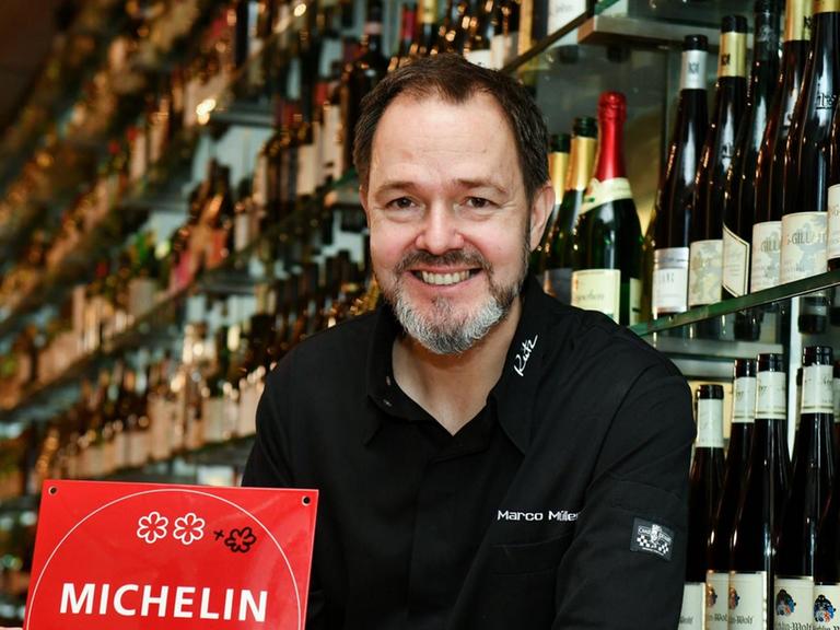Der Sternekoch Marco Müller in seinem Restaurant Rutz. Er hält einen Michelin-Restaurantführer in Händen und blickt lächelnd in Richtung des Betrachters.