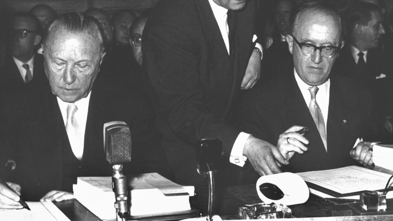 Das historische Schwarz-Weiß-Bild zeigt Adenauer und Hallstein nebeneinander an einem Tisch sitzend vor den Verträgen. Das historische Schwarz-Weiß-Bild zeigt Adenauer und Hallstein nebeneinander an einem Tisch sitzend vor den Verträgen.