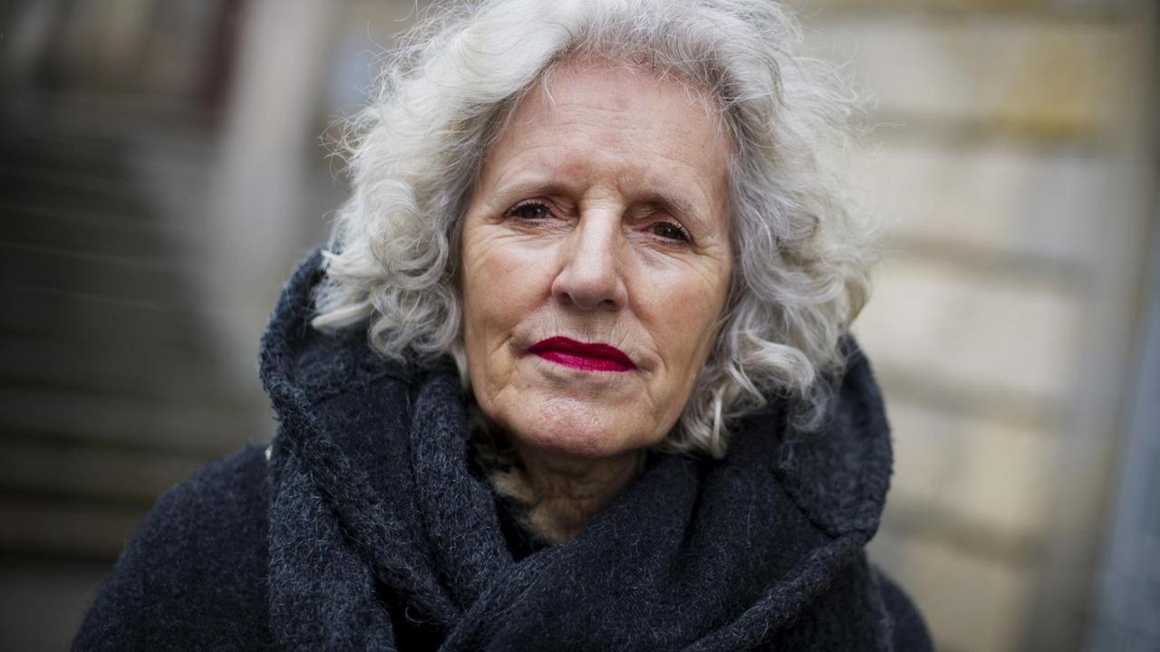 Nahaufnahme im Porträt: Ulrike Edschmid vor dem Hintergrund einer unscharen Fassade eines Gebäudes. Sie hat weiße Haare, trägt einen Mantel mit hohem Kragen und blickt ernst in die Kamera. Nahaufnahme im Porträt: Ulrike Edschmid vor dem Hintergrund einer unscharen Fassade eines Gebäudes. Sie hat weiße Haare, trägt einen Mantel mit hohem Kragen und blickt ernst in die Kamera.