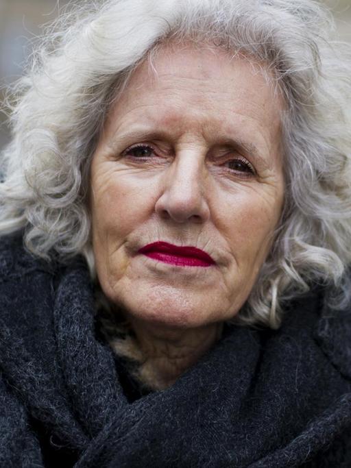 Nahaufnahme im Porträt: Ulrike Edschmid vor dem Hintergrund einer unscharen Fassade eines Gebäudes. Sie hat weiße Haare, trägt einen Mantel mit hohem Kragen und blickt ernst in die Kamera.