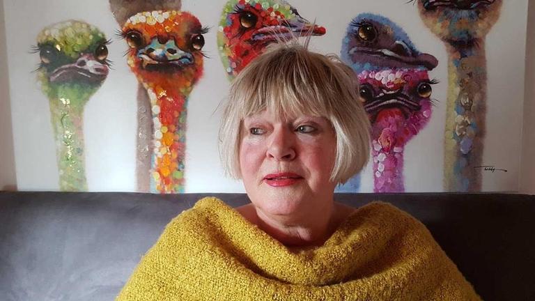 Eine Dame mit blondem Pagenkopf. Um die Schultern trägt sie einem senfgelben Schal. Im Hintergrund sieht man eine Tapete, von der bunte Vögel mit sehr langen Hälsen in den Raum schauen. Eine Dame mit blondem Pagenkopf. Um die Schultern trägt sie einem senfgelben Schal. Im Hintergrund sieht man eine Tapete, von der bunte Vögel mit sehr langen Hälsen in den Raum schauen.