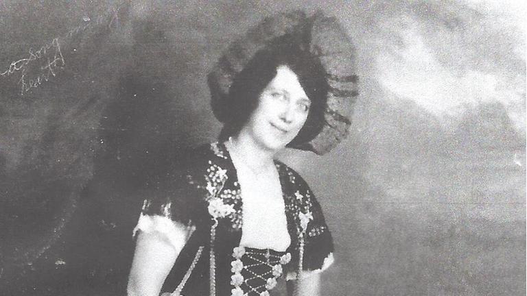 Eine junge Frau mit auffälligem Hut und Tracht lächelt auf einer historischen Fotografie in die Kamera.