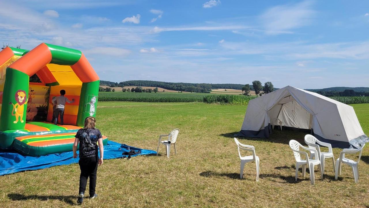 Eine Hüpfburg, ein weißes Zelt und weiße Plastikstühle stehen auf einer Wiese, im Hintergrund Hügel mit Wald und blauer Himmel. Eine Hüpfburg, ein weißes Zelt und weiße Plastikstühle stehen auf einer Wiese, im Hintergrund Hügel mit Wald und blauer Himmel.