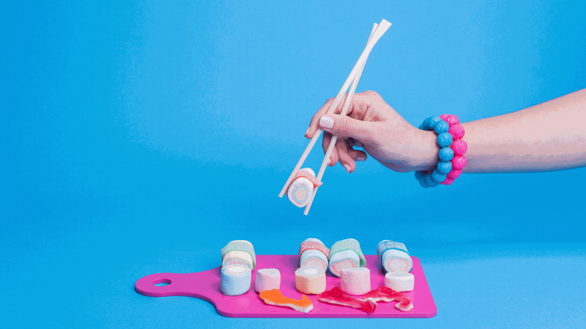 Marshmallows in Suhi-Form geschnitten, liegen auf einem rosafarbenen Brett. Eine Hand hält mit Stäbchen einen Marshmallow. Marshmallows in Suhi-Form geschnitten, liegen auf einem rosafarbenen Brett. Eine Hand hält mit Stäbchen einen Marshmallow.
