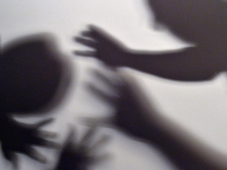 Schatten von Händen und zwei Menschen am Bildrand symbolisieren, wie eine Frau versucht, sich vor der Gewalt eines Mannes zu schützen.