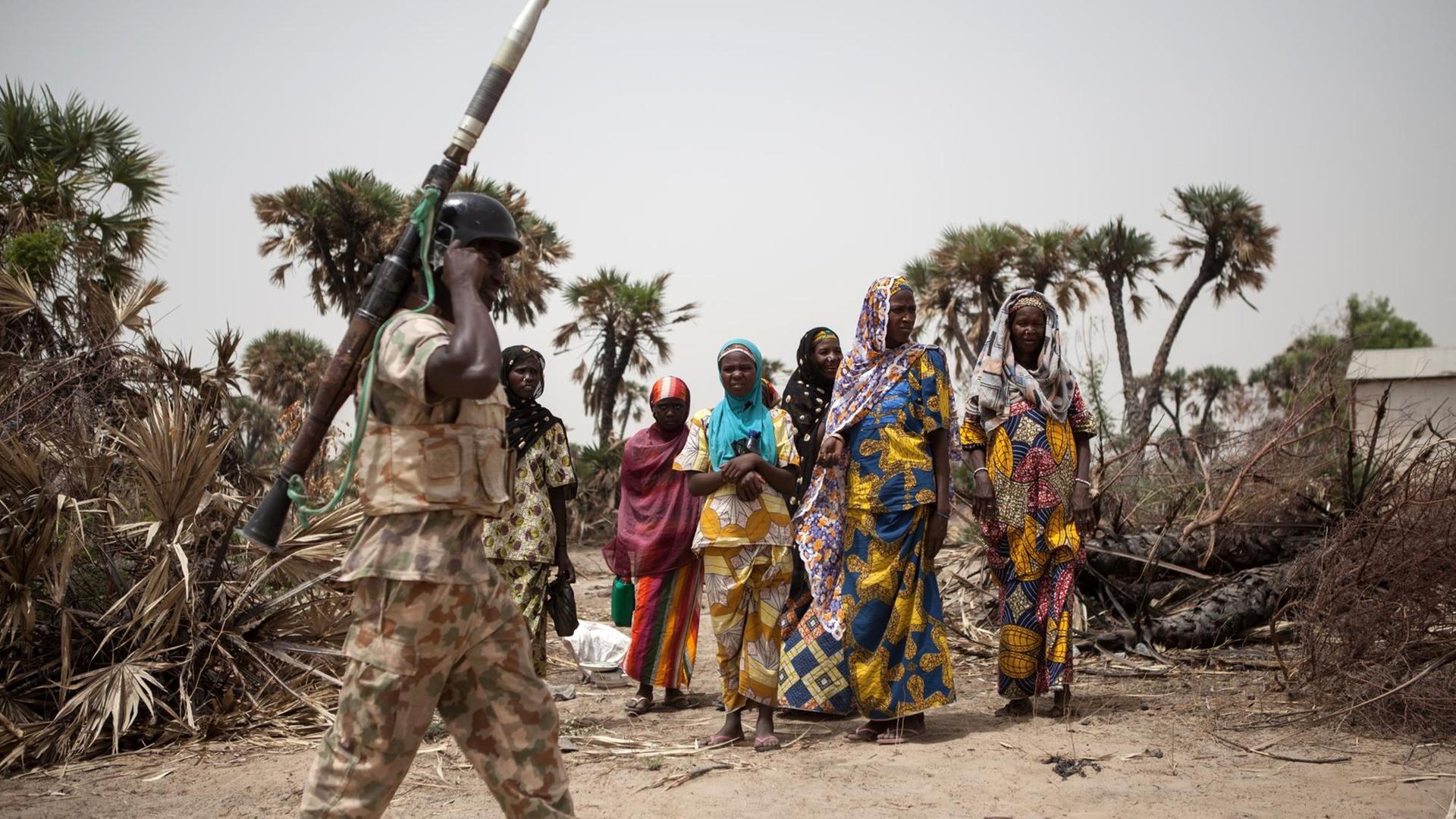 Soldat läuft mit einer reaktiven Panzerbüchse an der Schulter an einer Gruppe nigerianischer Frauen vorbei Soldat läuft mit einer reaktiven Panzerbüchse an der Schulter an einer Gruppe nigerianischer Frauen vorbei