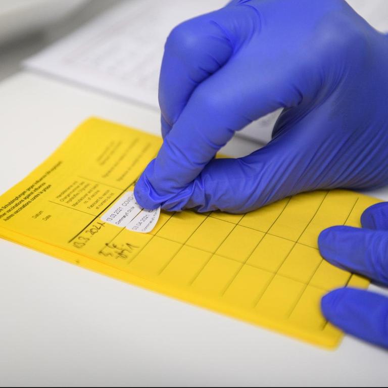 Eine Hand mit Schutzhandschuh klebt einen Aufkleber in einen gelben Impfpass 
