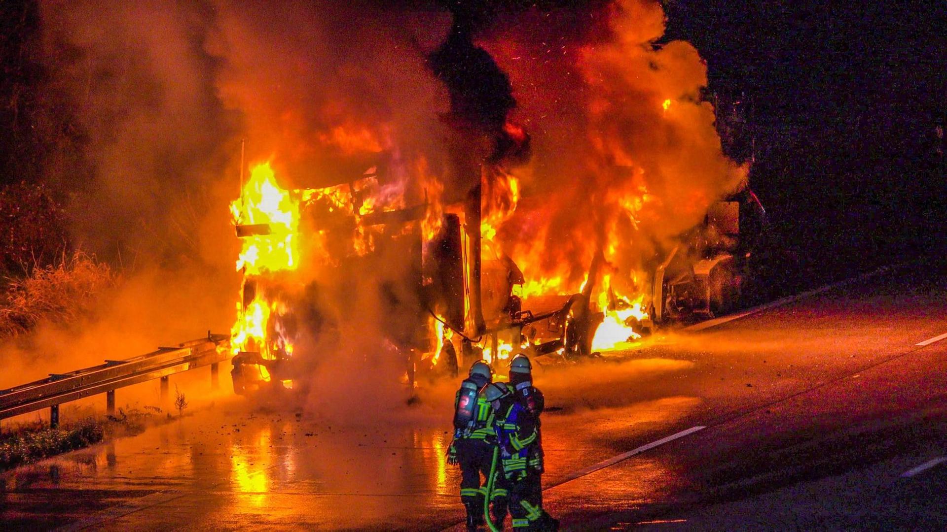 Feuerwehrleute stehen vor einem brennenden LKW, der mit Elektroautos beladen ist. Feuerwehrleute stehen vor einem brennenden LKW, der mit Elektroautos beladen ist.
