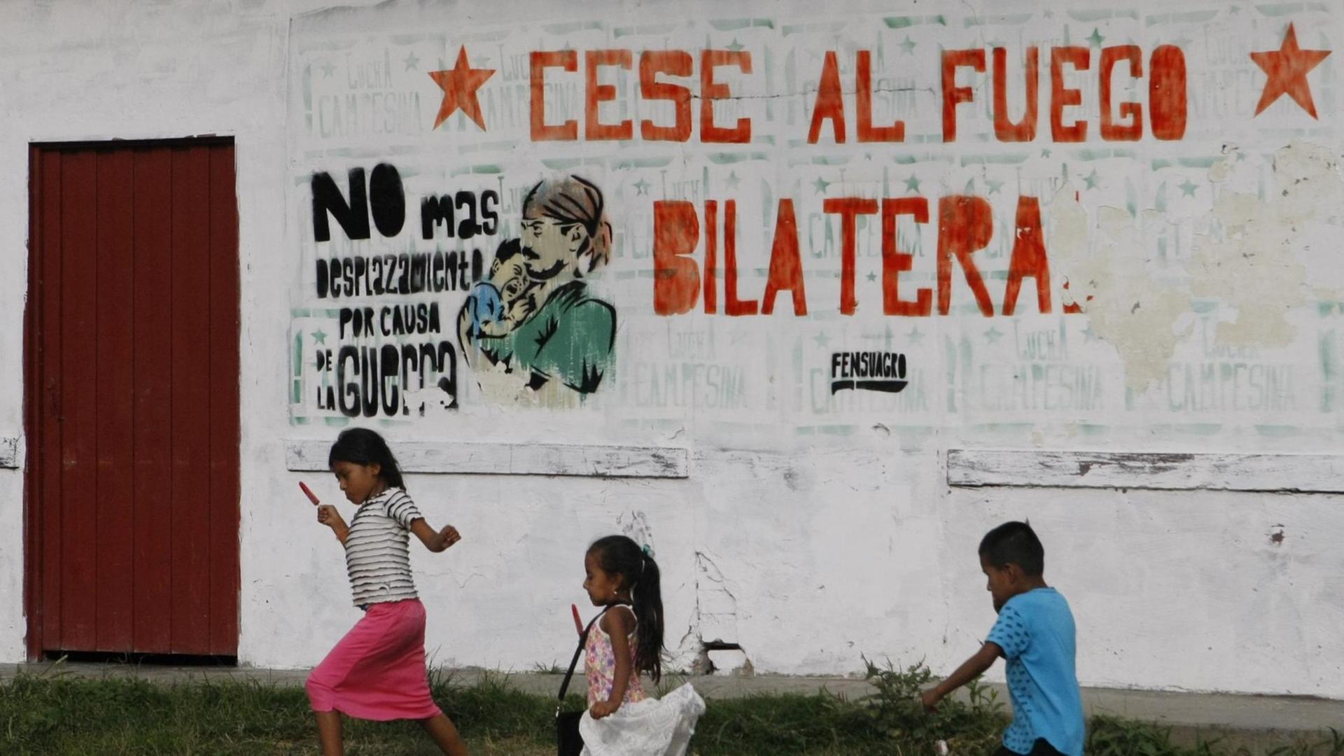 Kinder in Cauca spielen vor einer Mauer, die eine Aufschrift trägt ("Cese al fuego bilatera, "Bilaterale Feuerpause").