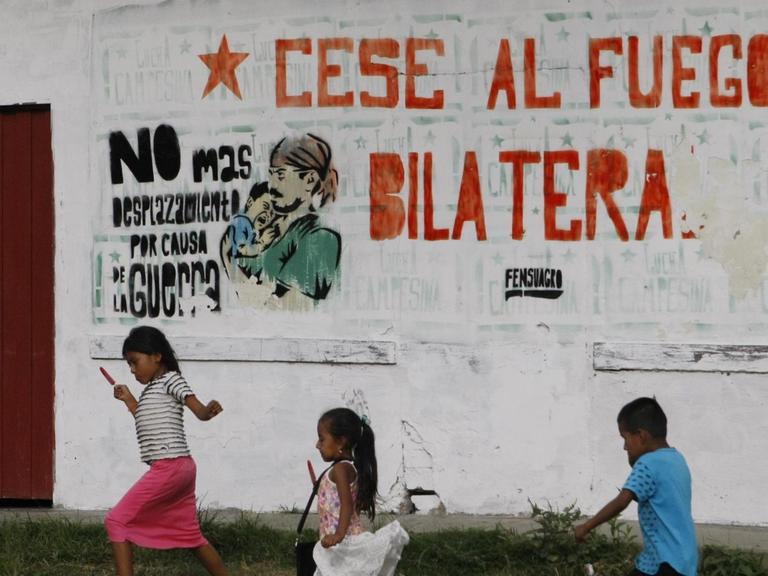 Kinder in Cauca spielen vor einer Mauer, die eine Aufschrift trägt ("Cese al fuego bilatera, "Bilaterale Feuerpause").