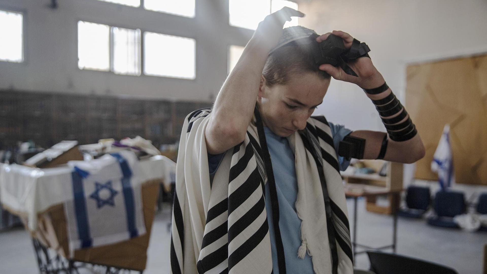 Ein Junge steht in einer großen Halle, im Hintergrund sind Fahnen mit Davidsternen zu erkennen, und befestigt gerade das Klötzchen des Tefillin am Kopf. Ein Junge steht in einer großen Halle, im Hintergrund sind Fahnen mit Davidsternen zu erkennen, und befestigt gerade das Klötzchen des Tefillin am Kopf.