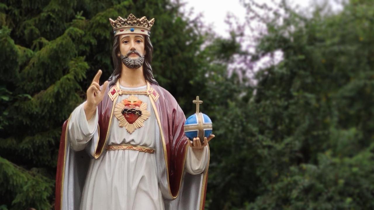 Eine lebensgroße Christusfigur mit rotem Umhang und goldener Krone hält die rechte Hand zu einer Segnugsgeste erhoben, sie steht im Freien auf einem Platz vor hohen Laub- und Nadelbäumen.