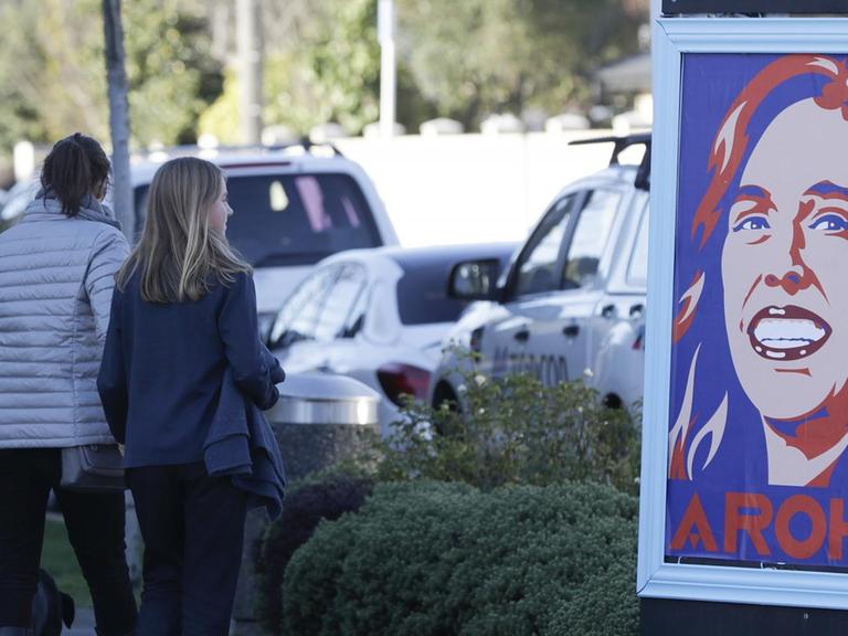 Straßenszene in Christchurch, Neuseeland: Zwei Fußgängerinnen und ein Hund passieren ein Plakat, dass die Premierministerin Jacinda Ardern mit dem Wort Aroha als Pop-Art-Illustration zeigt. Aroha bedeutet Liebe.