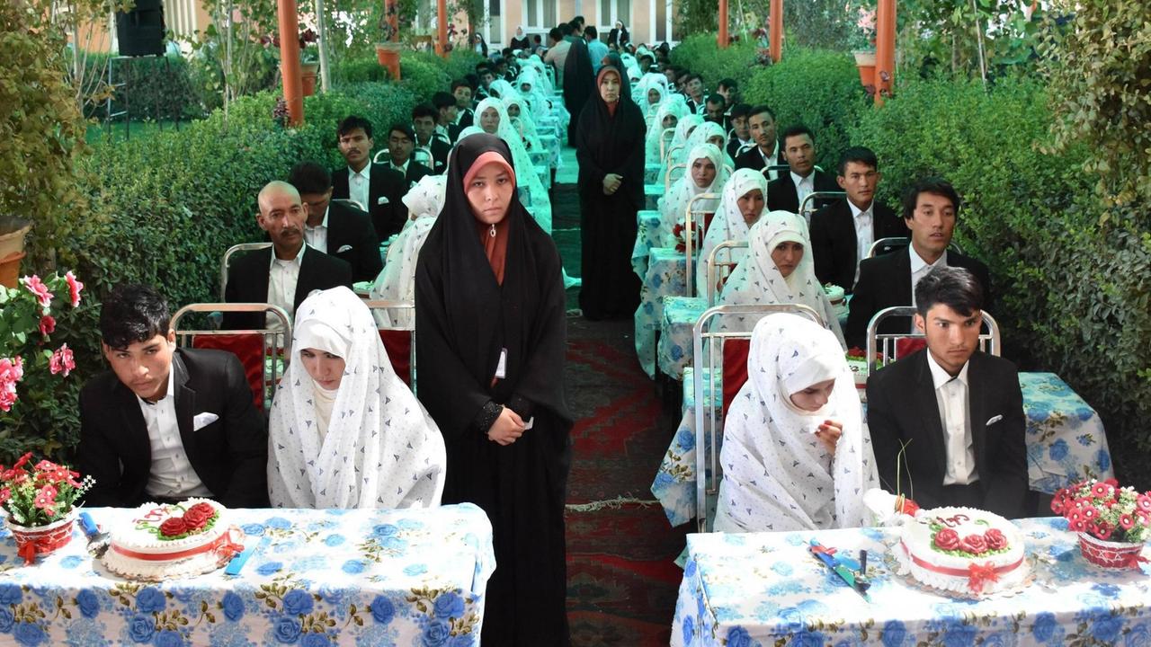 Gruppenhochzeit in Masar-e-Sharif in Afghanistan, aufgenommen im August 2018: Brautpaare - alle gleich gekleidet, die Männer in schwarzen Anzügen, die Frauen weiß verschleiert - sitzen an identisch dekorierten Tischen mit Hochzeitstorten hintereinander, und gucken ernst Richtung Kamera Gruppenhochzeit in Masar-e-Sharif in Afghanistan, aufgenommen im August 2018: Brautpaare - alle gleich gekleidet, die Männer in schwarzen Anzügen, die Frauen weiß verschleiert - sitzen an identisch dekorierten Tischen mit Hochzeitstorten hintereinander, und gucken ernst Richtung Kamera