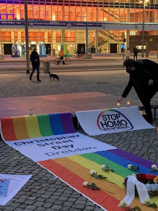 Eine Regenbogenfahne, ein Kranz und weiße Blumen erinnern auf dem Altmarkt vor dem Kulturpalast in Dresden an die Opfer einer Messerattacke