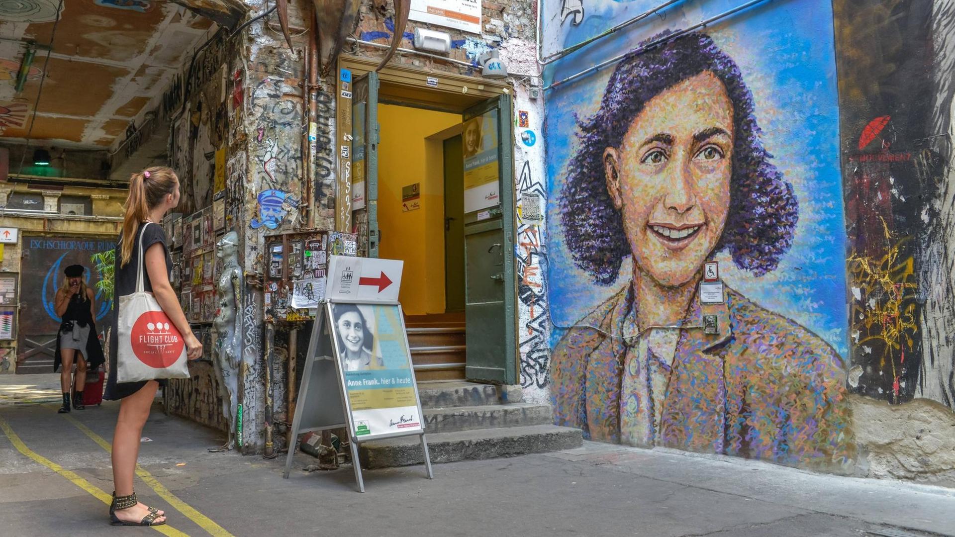 Ein junge Frau mit Stoffbeutel vor einem Graffiti, das Anne Frank zeigt, vor dem Anne Frank Zentrum im Hinterhof an der Rosenthaler Straße in Berlin-Mitte Ein junge Frau mit Stoffbeutel vor einem Graffiti, das Anne Frank zeigt, vor dem Anne Frank Zentrum im Hinterhof an der Rosenthaler Straße in Berlin-Mitte