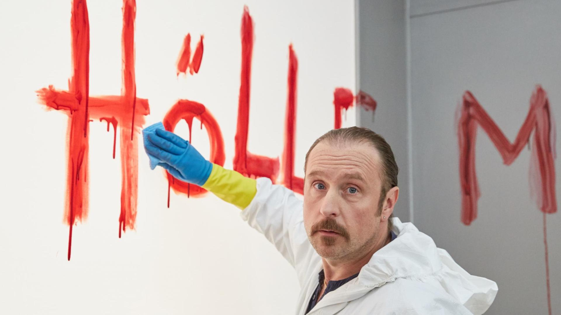 Der Schauspieler Bjarne Mädel trägt einen weißen Anzug und schrubbt das mit Blut an die Wand geschriebene Wort "Hölle"