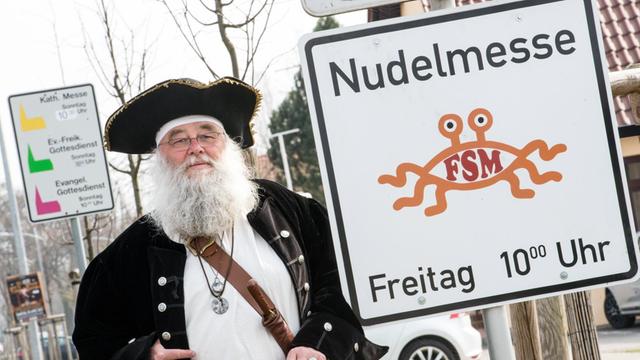 Rüdiger Weida von der "Kirche des Fliegenden Spaghettimonsters" steht am 05.04.2016 neben seinem Schild "Nudelmesse" in Templin (Brandenburg) und im Hintergrund ist das Schild der katholischen und evangelischen Kirche zu sehen.