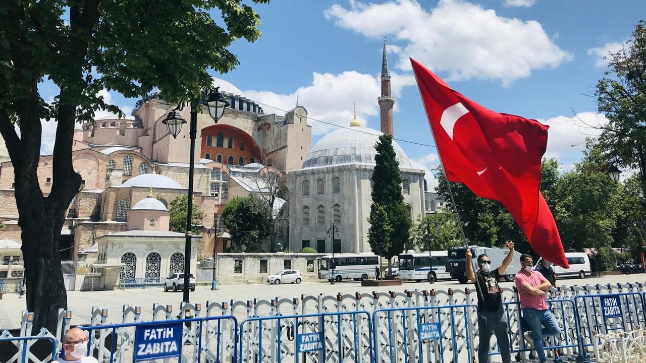 Ein Mann mit Mundschutz steht vor den Absperrungen vor der Hagia Sophia und schwenkt die rote Fahne der Türkei. Ein Mann mit Mundschutz steht vor den Absperrungen vor der Hagia Sophia und schwenkt die rote Fahne der Türkei.