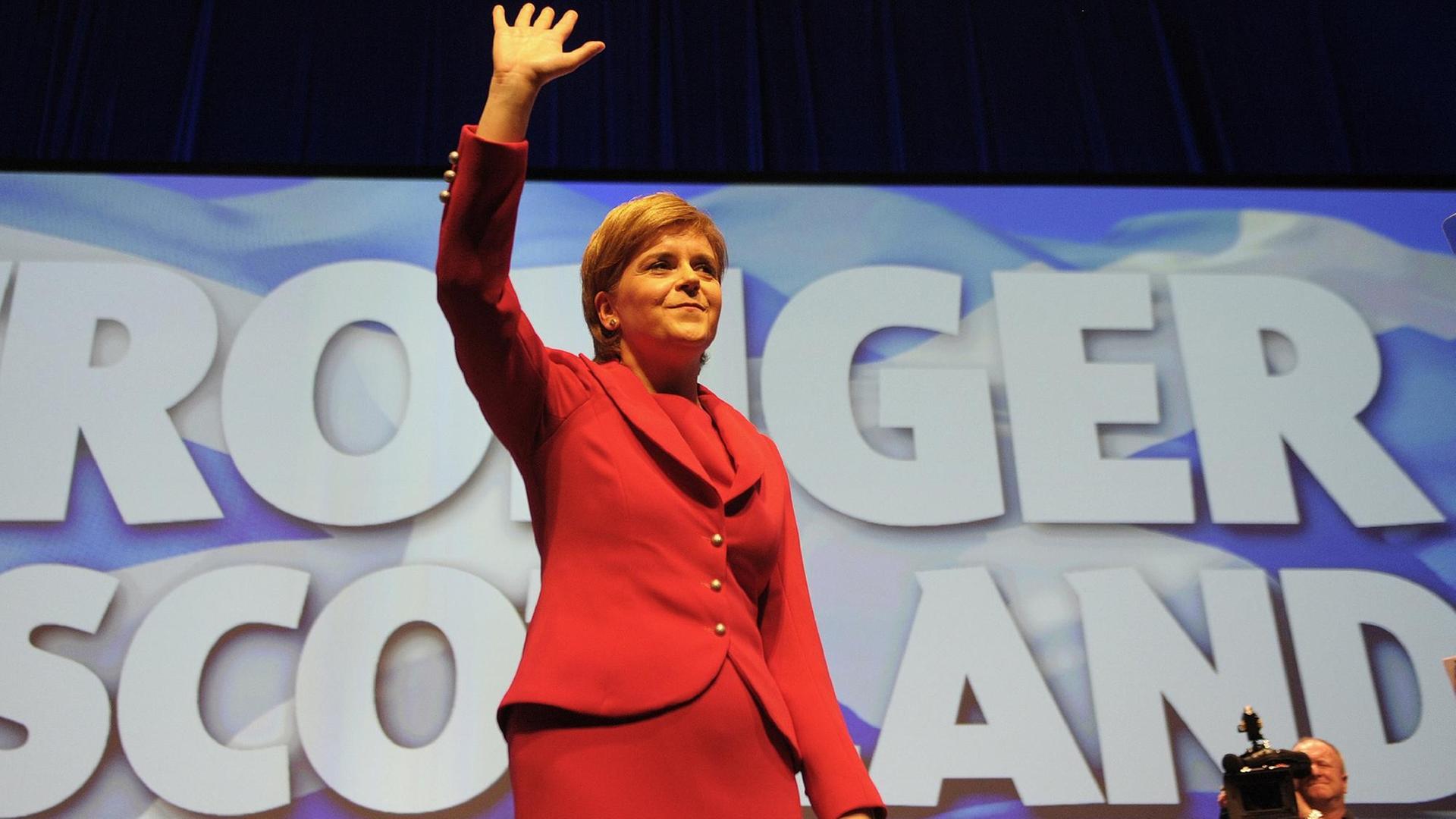 Das Bild zeigt Schottlands Regierungschefin Nicola Sturgeon auf dem SNP-Parteitag am 15.10.2016 in Glasgow. Sie trägt ein rotes Kleid und winkt mit der rechten Hand den Delegierten zu. Hinter ihr geschrieben steht "Stronger Scotland - ein stärkerers Schottland".