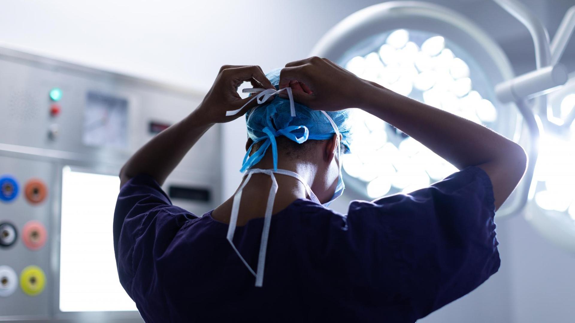 Eine Chirurgin ist im Operationssaal eines Krankenhauses von hinten zu sehen, während sie sich eine OP-Maske zubindet. Eine Chirurgin ist im Operationssaal eines Krankenhauses von hinten zu sehen, während sie sich eine OP-Maske zubindet.
