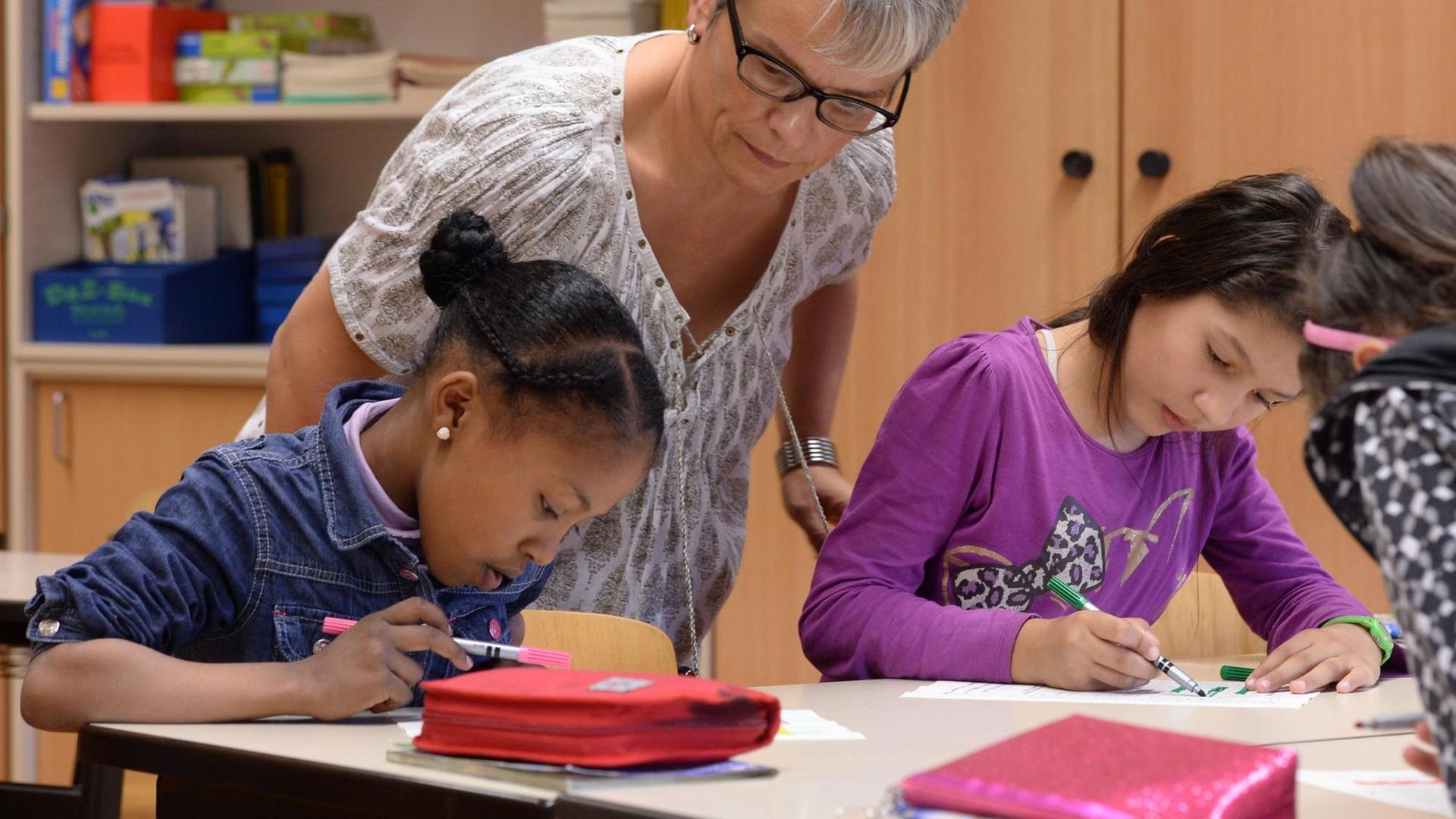 Zwei Kinder beugen sich über einen Schreibtisch und notieren etwas, eine Lehrerin steht hinter ihnen und beugt sich über sie. Zwei Kinder beugen sich über einen Schreibtisch und notieren etwas, eine Lehrerin steht hinter ihnen und beugt sich über sie.