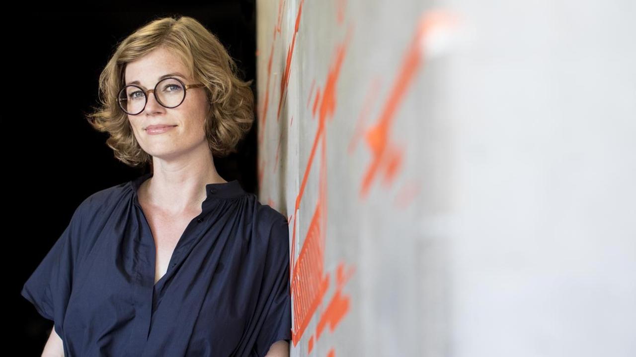 Porträt der Unternehmerin Insa Klasing, sie trägt eine runde Brille und ein blaues Kleid und sie lehnt an einer Wand in in der Factory Berlin, aufgenommen 2018 Porträt der Unternehmerin Insa Klasing, sie trägt eine runde Brille und ein blaues Kleid und sie lehnt an einer Wand in in der Factory Berlin, aufgenommen 2018