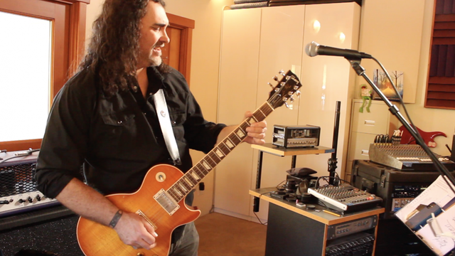 Ein Mann mit schwarzen langen Haaren steht in einem Studio vor einem Mikro und spielt E-Gitarre.