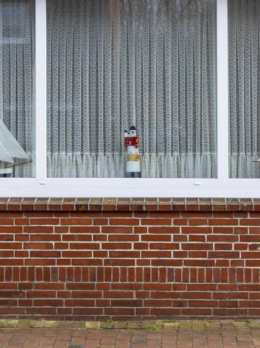 Fassade eines Backsteinhauses. Im Fenster steht ein Leuchtturm und zwei Segelboote, die Gardinen sind zugezogen.