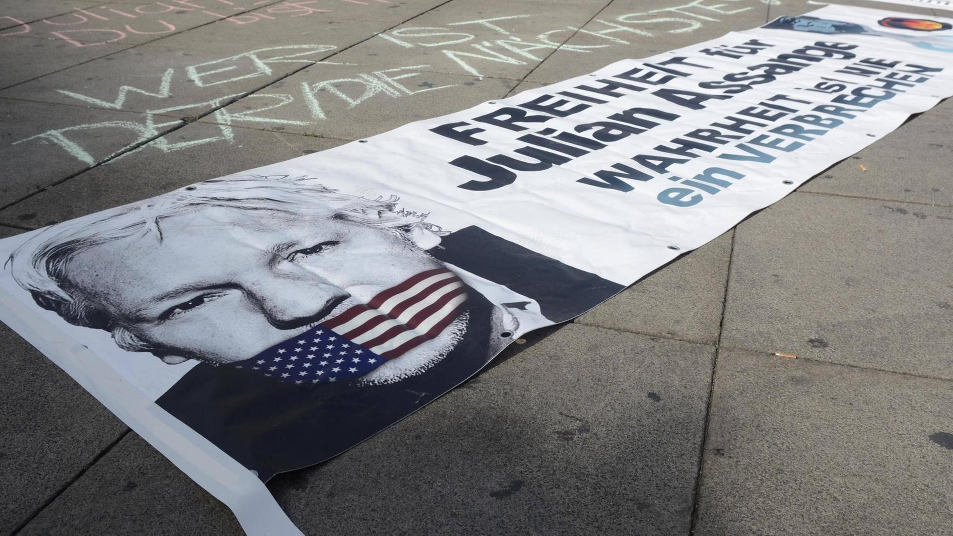 Auf einem Plakat, das auf der Straße liegt, ist das Bild von Julian Assange zu sehen mit einem Maulkorb in den US-Farben. Auf einem Plakat, das auf der Straße liegt, ist das Bild von Julian Assange zu sehen mit einem Maulkorb in den US-Farben.