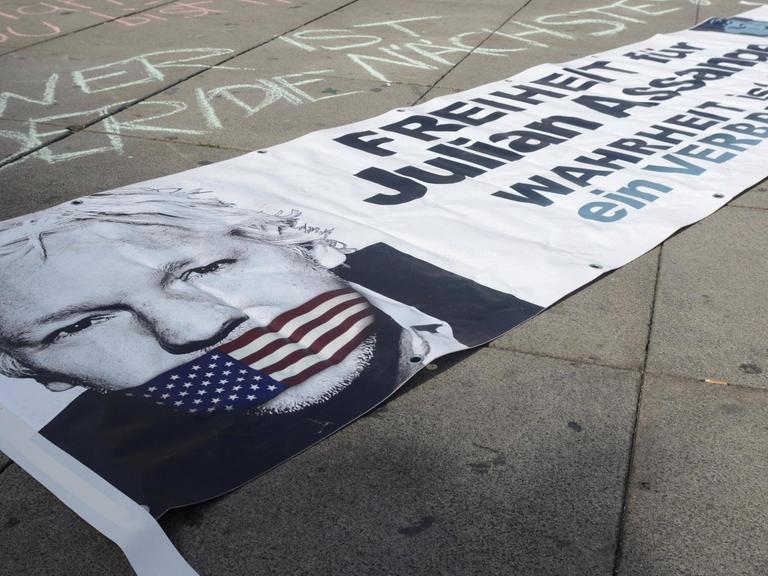 Auf einem Plakat, das auf der Straße liegt, ist das Bild von Julian Assange zu sehen mit einem Maulkorb in den US-Farben.